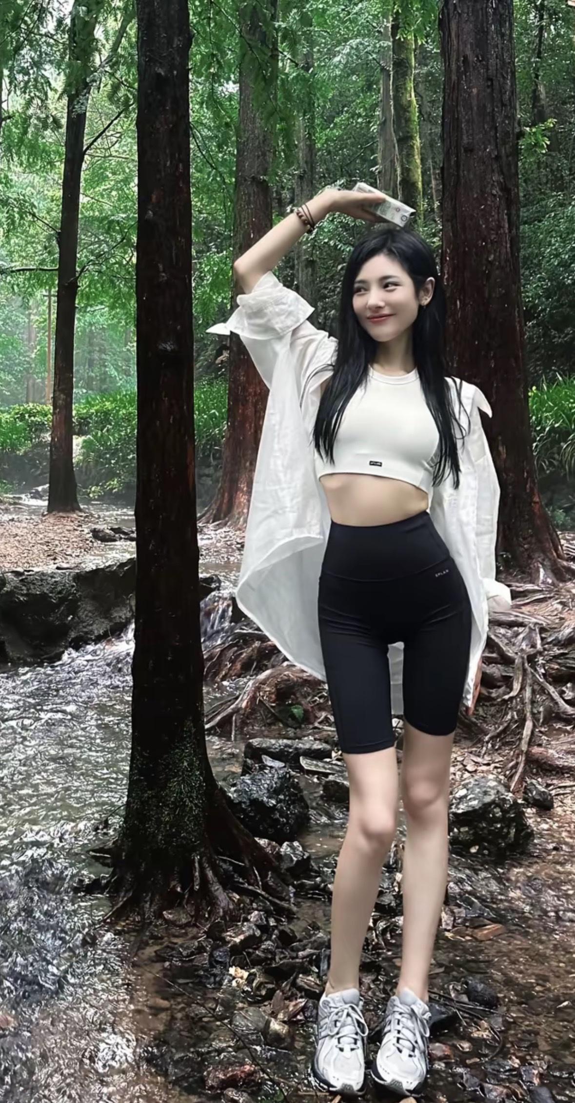 林间溪畔，绿意盎然。女孩身着白色短上衣与黑色骑行裤，外搭宽松白衬衫，脚踩运动鞋，