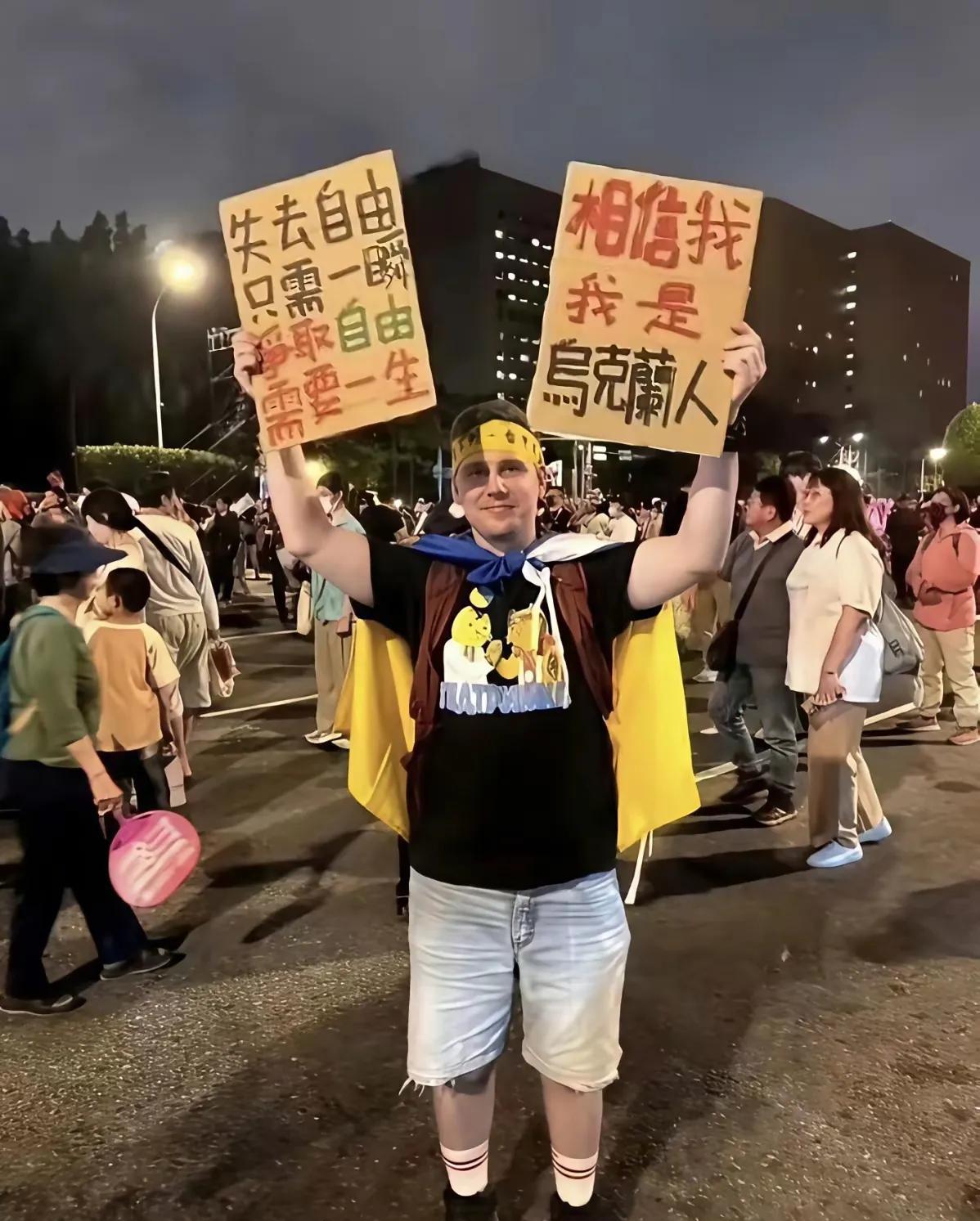 一位身在异国他乡的乌克兰人，身披乌克兰国旗，高举着两块牌子。一块上面用中文写着：