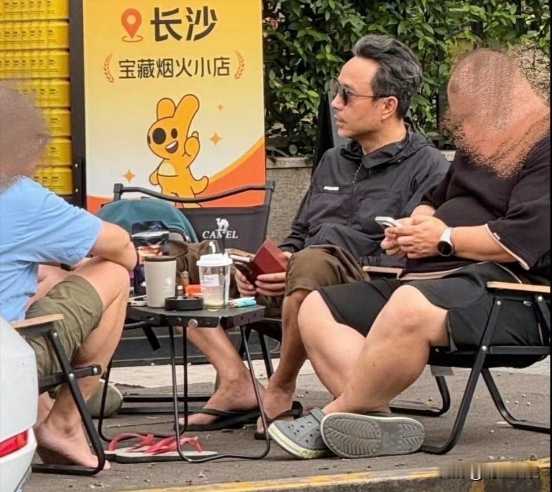 汪涵在街头坐着和朋友聊天，穿着人字拖吃着槟榔，用着三折叠手机，头发两侧都白了，但