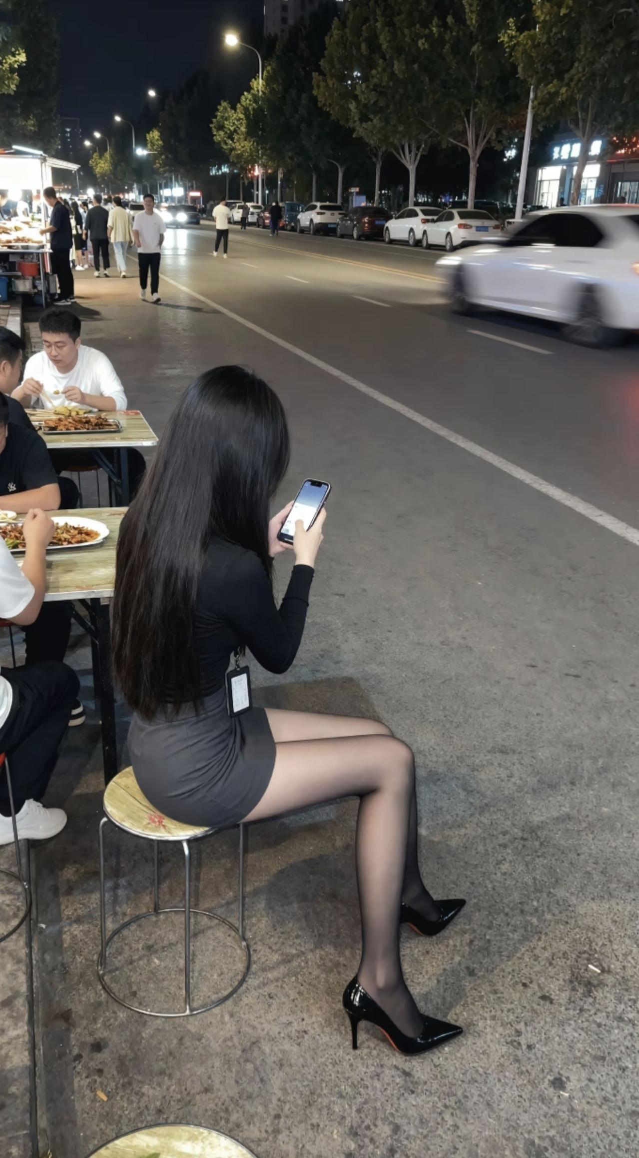 夜晚的路边摊，烟火气裹着晚风。长黑发配黑裙，高跟鞋踩着市井的热闹，手机屏亮着，是