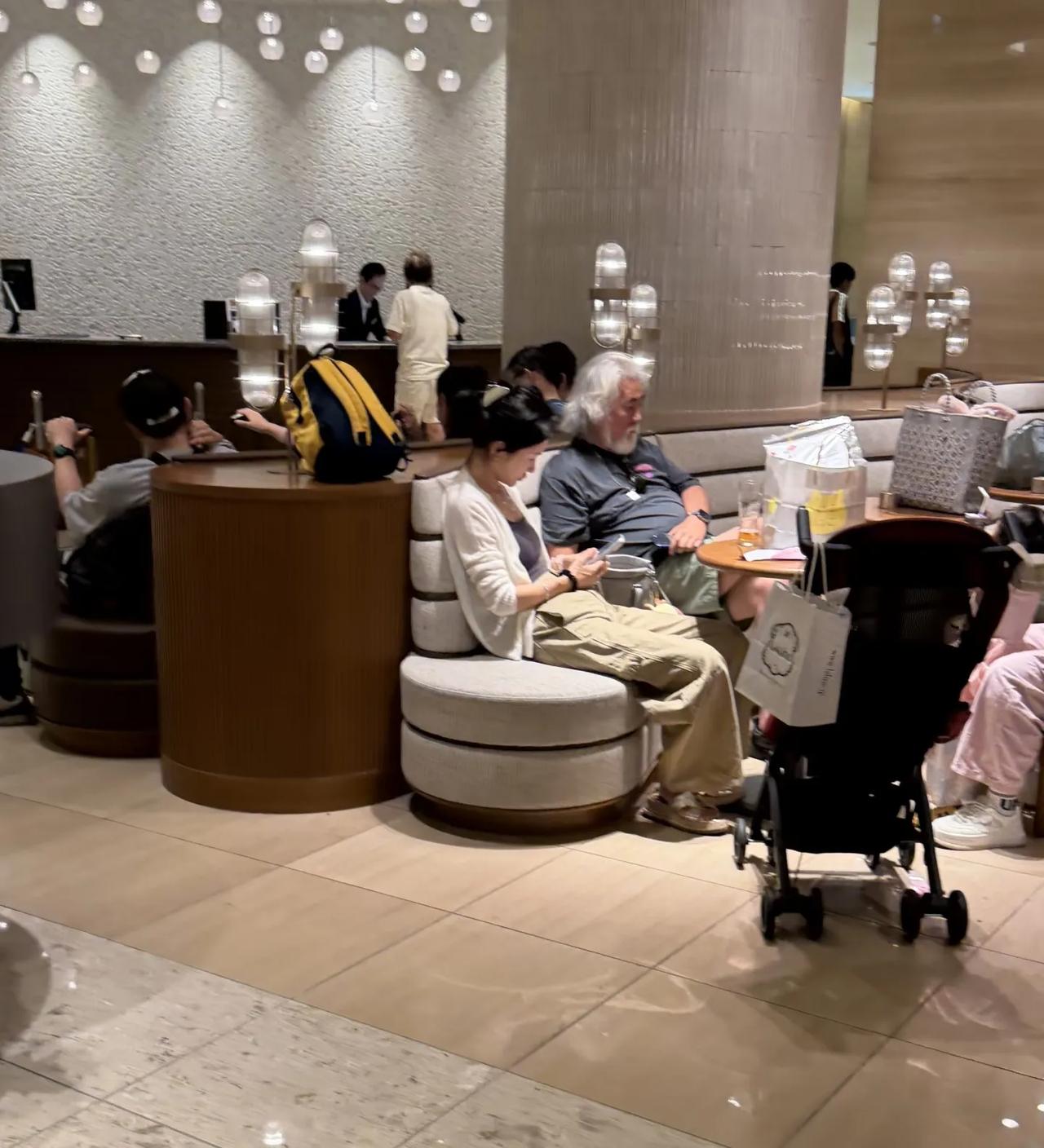 张纪中与妻子杜星霖坐在东京迪士尼希尔顿酒店大厅，杜星霖低头玩着手机，一旁的张