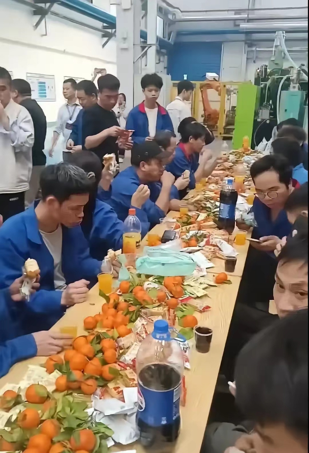 比亚迪长沙工厂开年会，车间里摆了几十桌，工人围坐吃盒饭。台上领导讲话，台下工人低