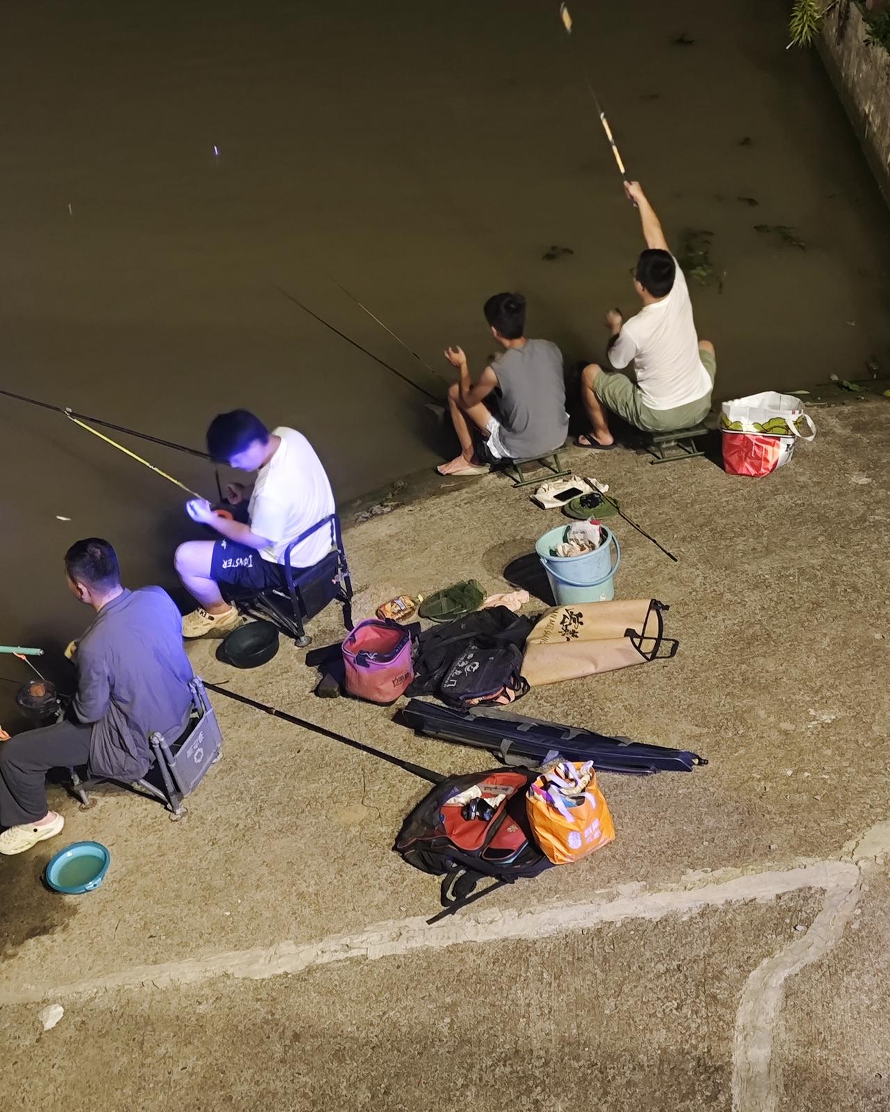 夜晚河边夜钓，几位钓友排坐岸边，鱼竿一字排开，夜色下氛围感拉满。岸边放着装