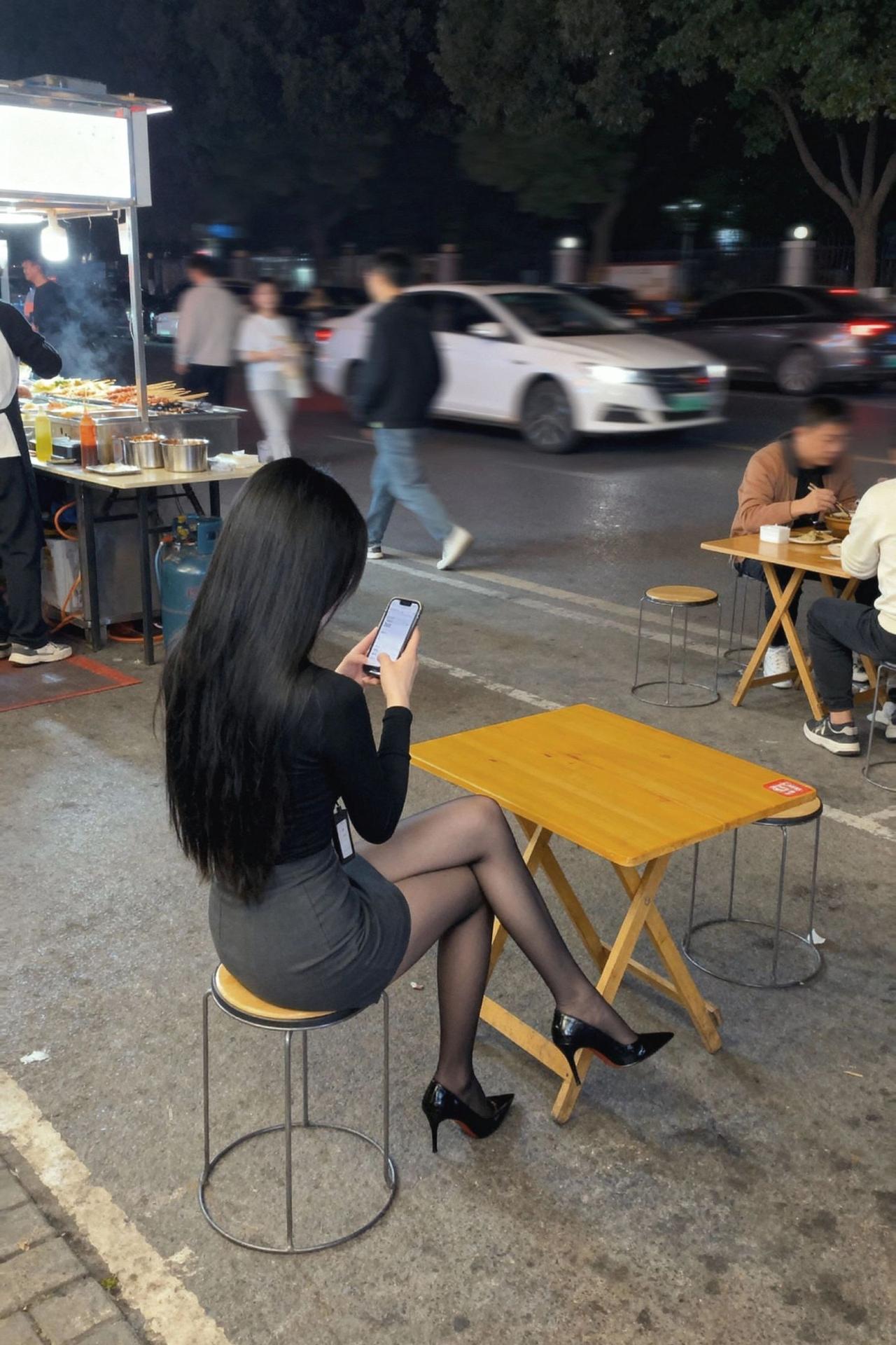 夜晚街边，时尚女孩低头玩手机，高跟鞋与长裙相得益彰，都市生活的一隅。