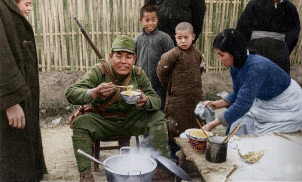 1937年12月的南京，一名日本兵背着三八式步枪、戴着军帽，坐在小板凳上，双手端