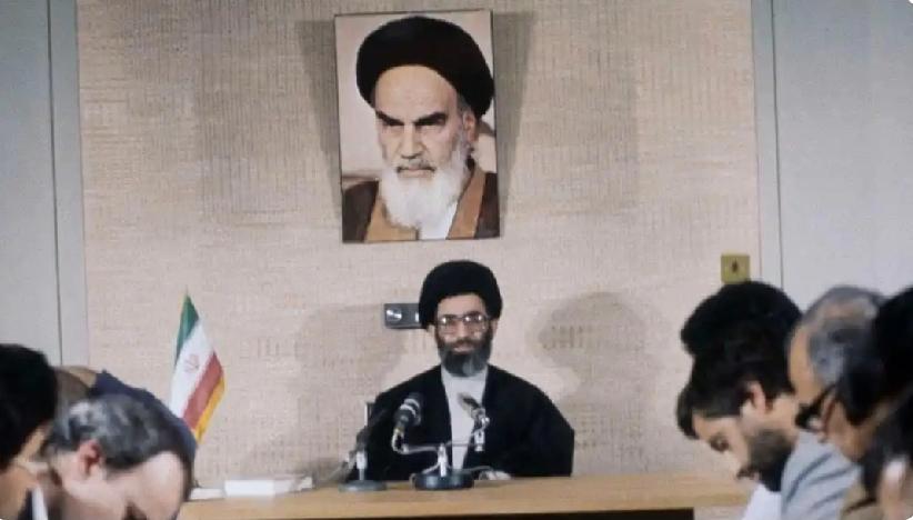 哈梅内伊端坐中央，正在开会。他身后的墙上挂着霍梅尼的照片。哈梅内伊（最左边）