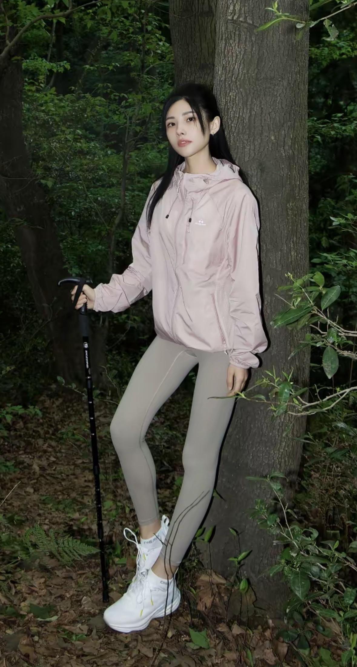 少女倚树而立，浅粉冲锋衣搭配灰调瑜伽裤，手持登山杖，脚踩白色运动鞋。夜色中的林间