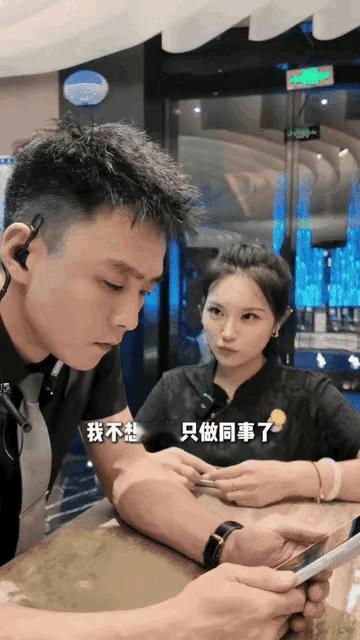 帅气男同事在桌上看着手机，女同事坐在他旁边摆弄着双手娇声娇气地说，我不想和你做同