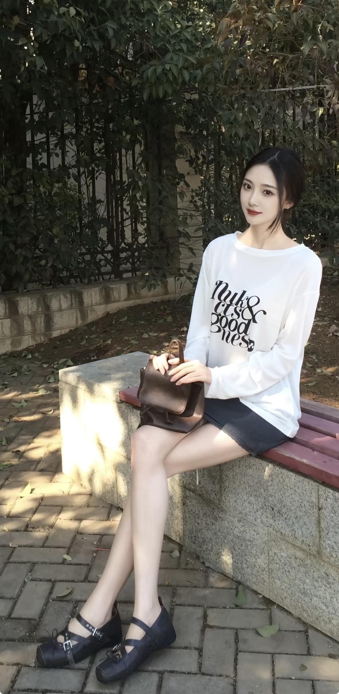 一位年轻女子坐在户外石凳上，身穿白色字母长袖T恤搭配深色短裤，脚踩黑色玛丽珍鞋，