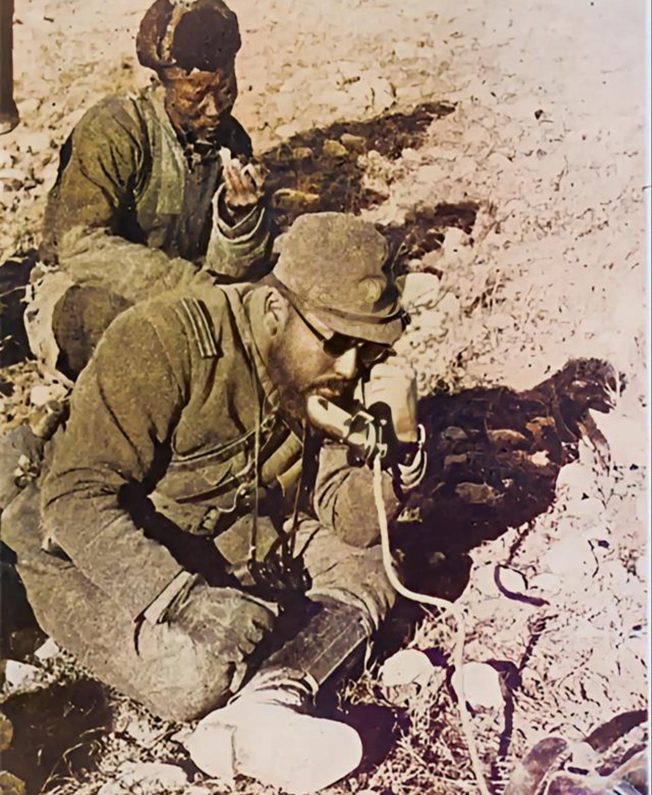 画面里，前景是一名留着胡须、戴着墨镜的侵华日军士官，他正蹲在荒地上，操作一台老式