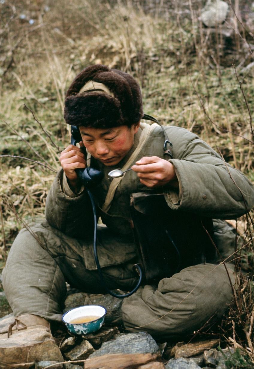 抗美援朝战争，年轻的志愿军通信兵身着厚实防寒棉服、头戴棉军帽，蹲踞在朝鲜前线的野