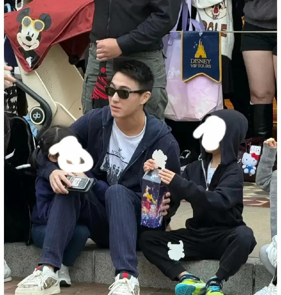 何猷君一个人背着双肩包，左手牵儿子右手抱女儿，在香港迪士尼排队买爆米花，照片里笑