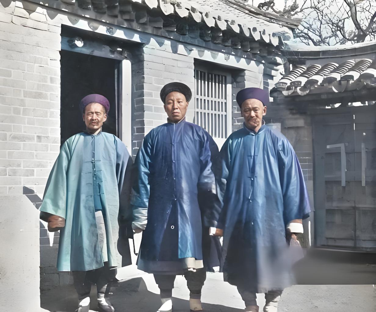 这是20世纪初的老照片，定格三位穿长袍、戴传统帽饰的村董，他们站在中式砖瓦房前。