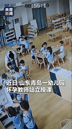 “被央媒点赞了！”山东青岛，一怀孕的幼儿园老师站着给小朋友授课，旁边桌的一小女孩