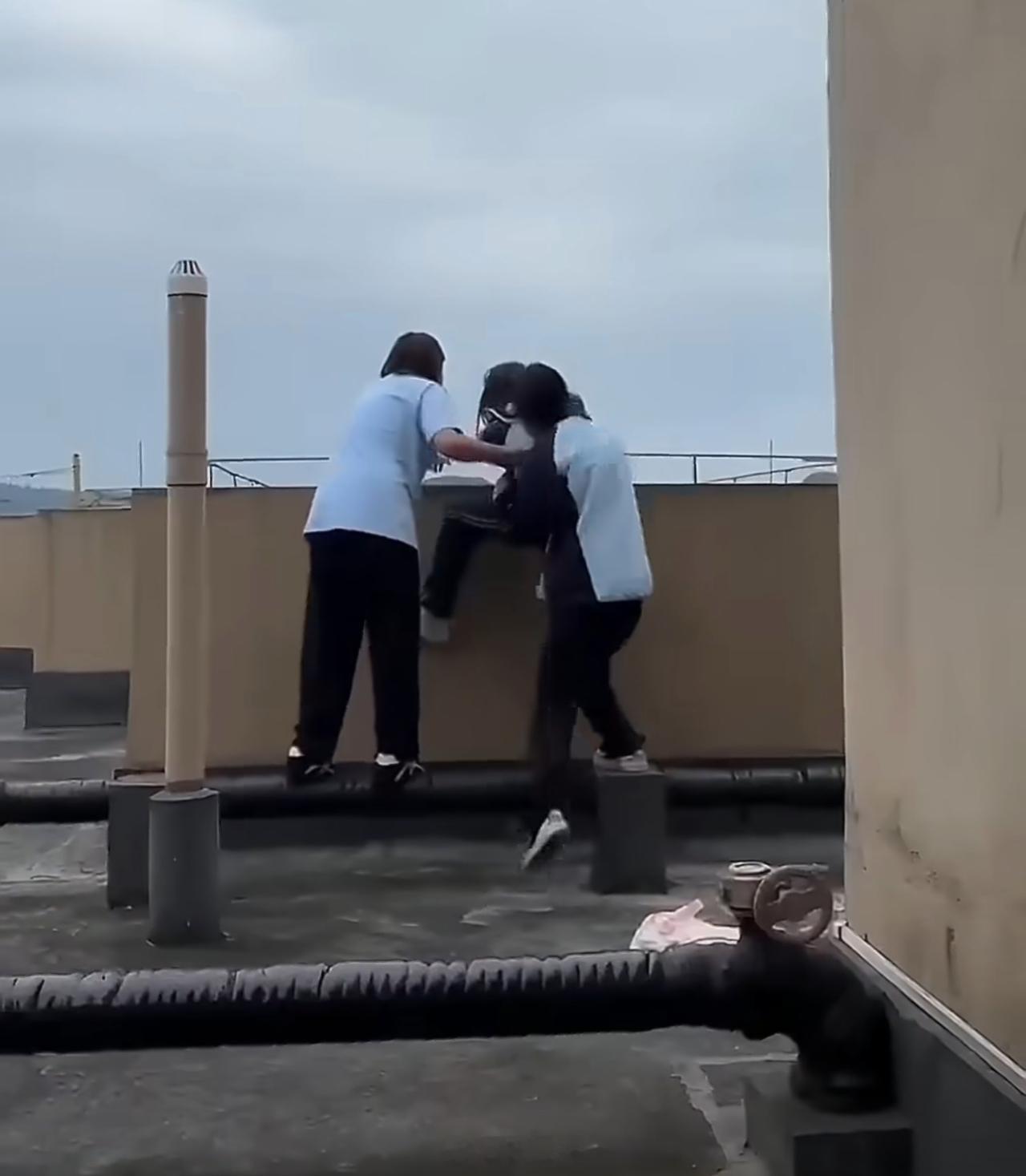 楼顶边缘，一名女生死死抓住栏杆，尖叫。两名体型更大的女生将她往女儿墙方向推。老师赶到，制止。这段画面