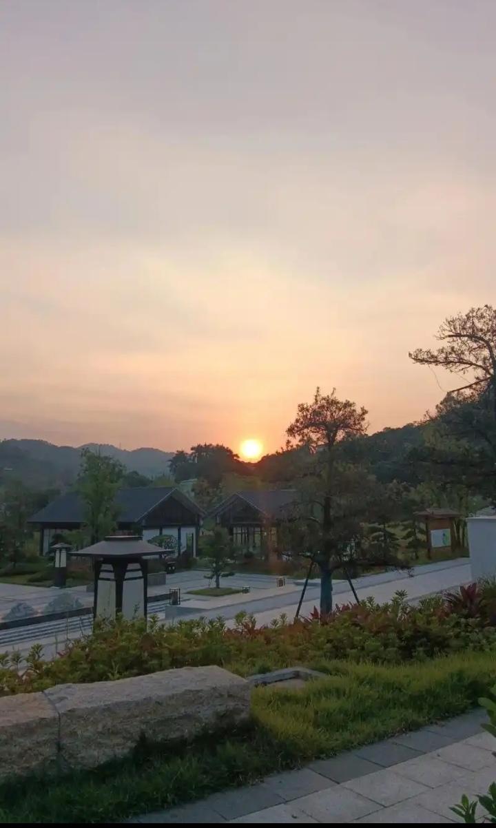 暮色轻笼庭院，落日将天边晕染成暖橙。黛瓦小屋隐在树影间，石阶旁草木舒展，远山如淡