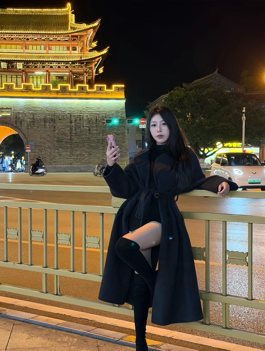 夜晚的古城墙下，黑衣少女自拍，手机亮起屏幕，背景灯火辉煌。
