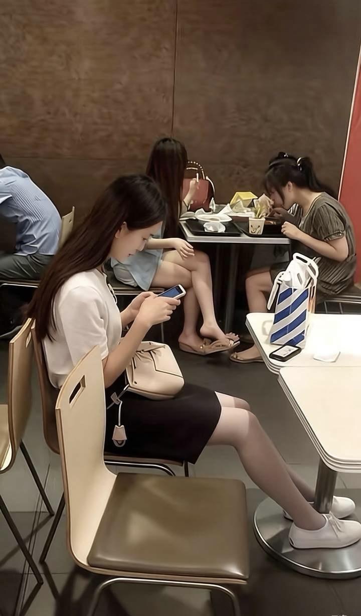 快餐店里，暖光轻洒。前景女子穿白上衣、黑短裙与灰丝袜，坐米色椅上，专注盯手机，浅