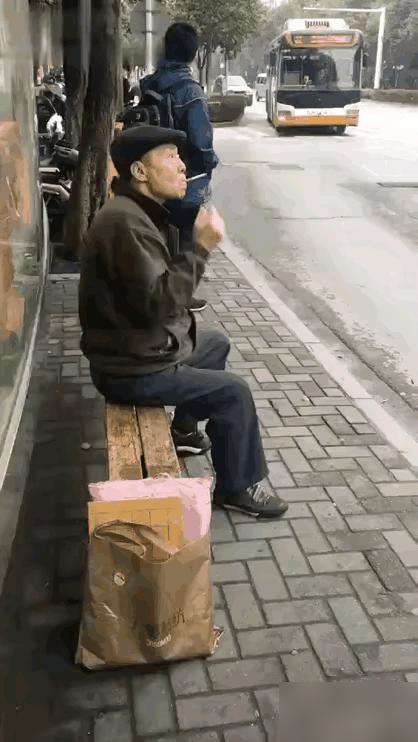 一名戴着帽子的大爷，坐在公交车站台等公交车。大爷等车的过程中，拿出一根烟，点燃后