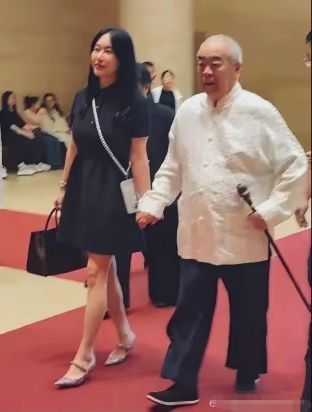当代著名书画家范曾携夫人徐萌走红毯。范曾身着中式白色对襟上衣，拄着手杖，气度沉稳