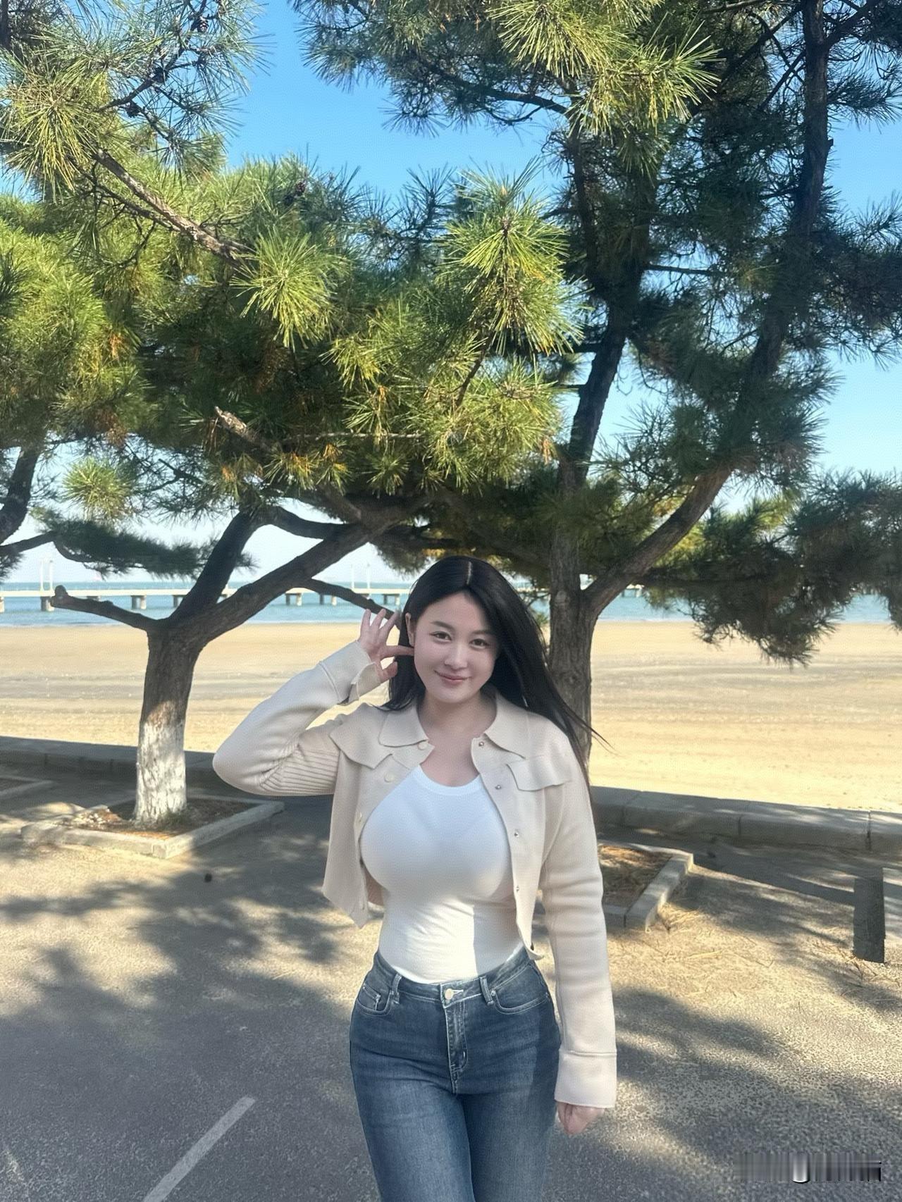 在阳光灿烂的海边，一位美女在松树旁驻足留影。她那比阳光还要夺目的笑容，温暖至极。