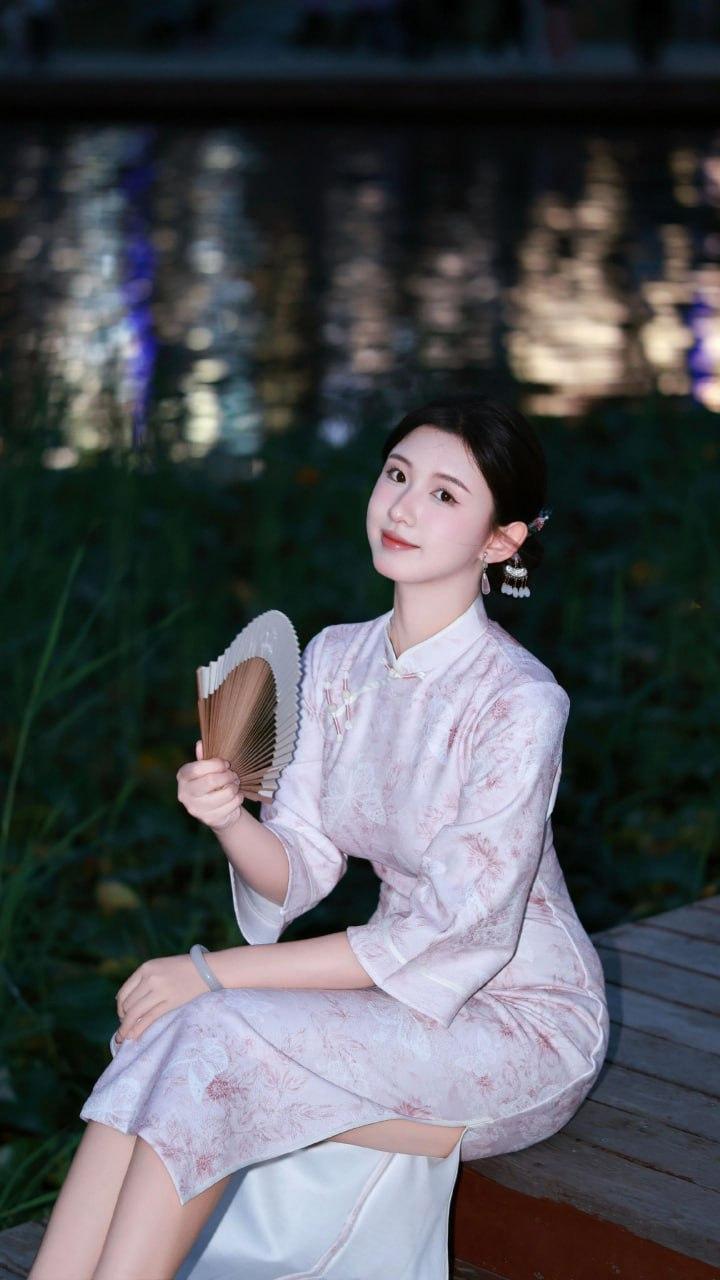 夜幕下的湖畔，一位女子身着淡粉印花旗袍，优雅坐在木栈道上。她发髻轻挽，缀着精致