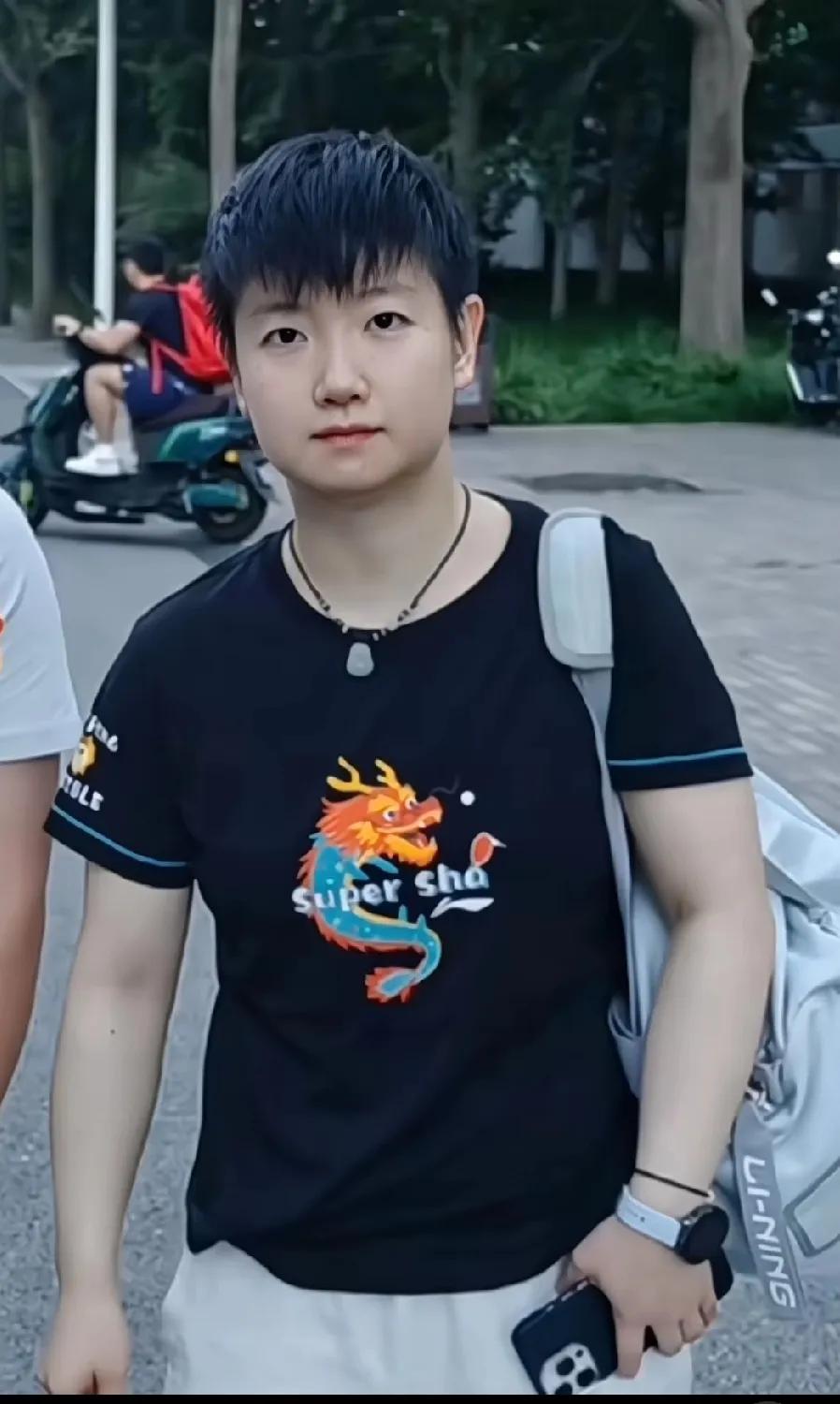孙颖莎背着单肩包，手里拿着手机，穿黑色印小龙图案的衣服，青春又可爱~赛场外的她