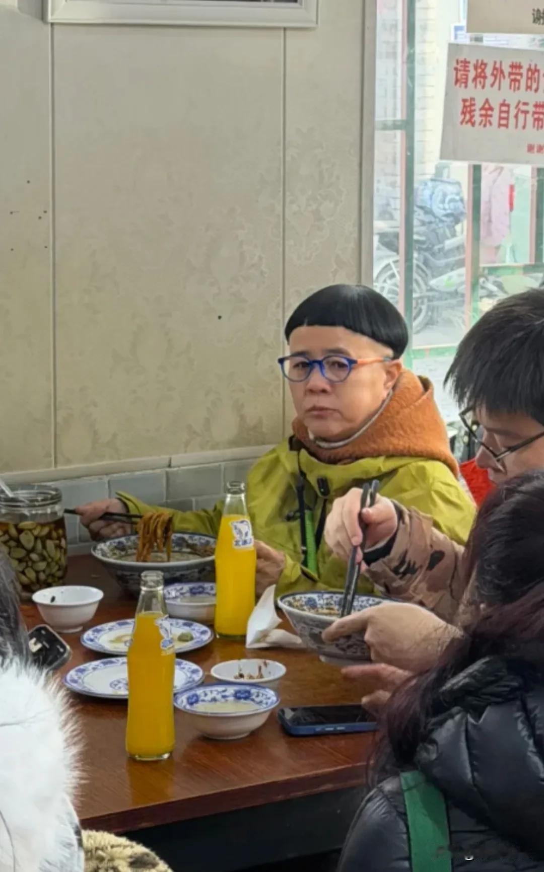金龟子老师在一间面馆吃炸酱面，留着娃娃头，但是头发挺乌黑浓密的，戴着黑框眼镜，穿