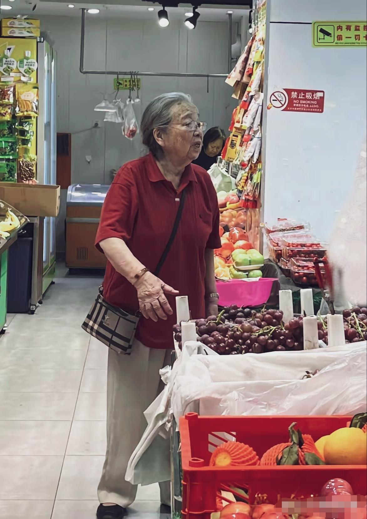 90岁李明启老师逛水果店，走路缓慢，背着一小包，穿搭简单朴素，与普通老奶奶一样接