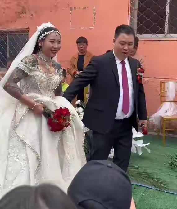 这是一场农村婚礼，新娘穿着满是珍珠和蕾丝的白色婚纱，手臂上有大面积纹身，手里捧着