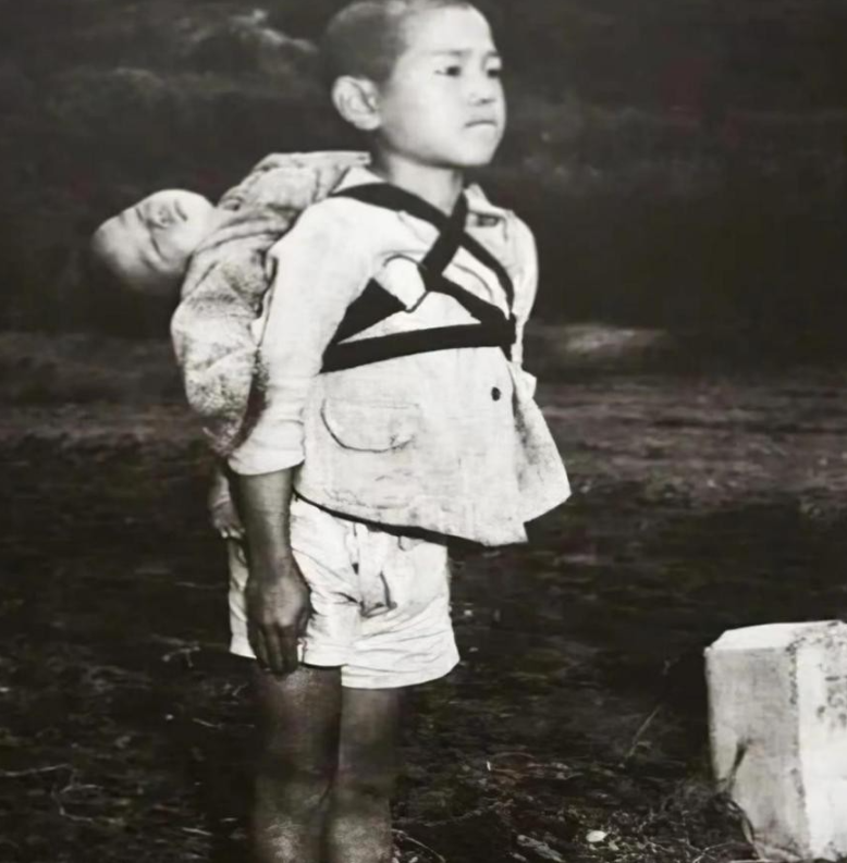1945年，一名10岁的日本男孩抿着嘴，笔直地背着弟弟。而弟弟头向后仰，早已气绝