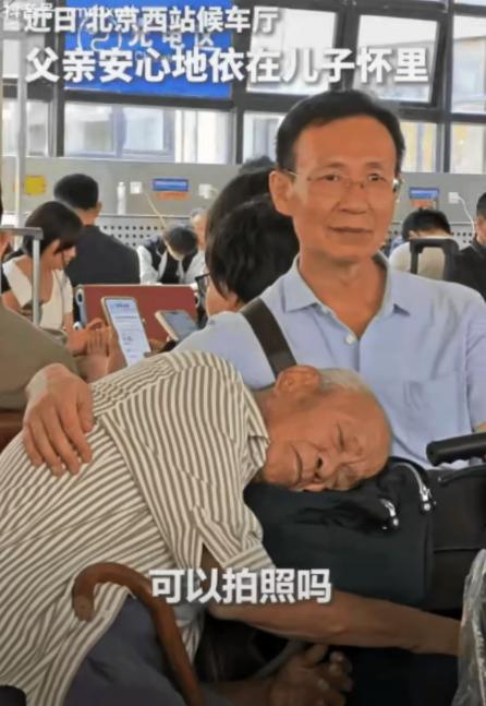 “火到人民日报了！”北京候车厅，一老人依偎在儿子怀里睡觉。一女乘客问：“拍个照可