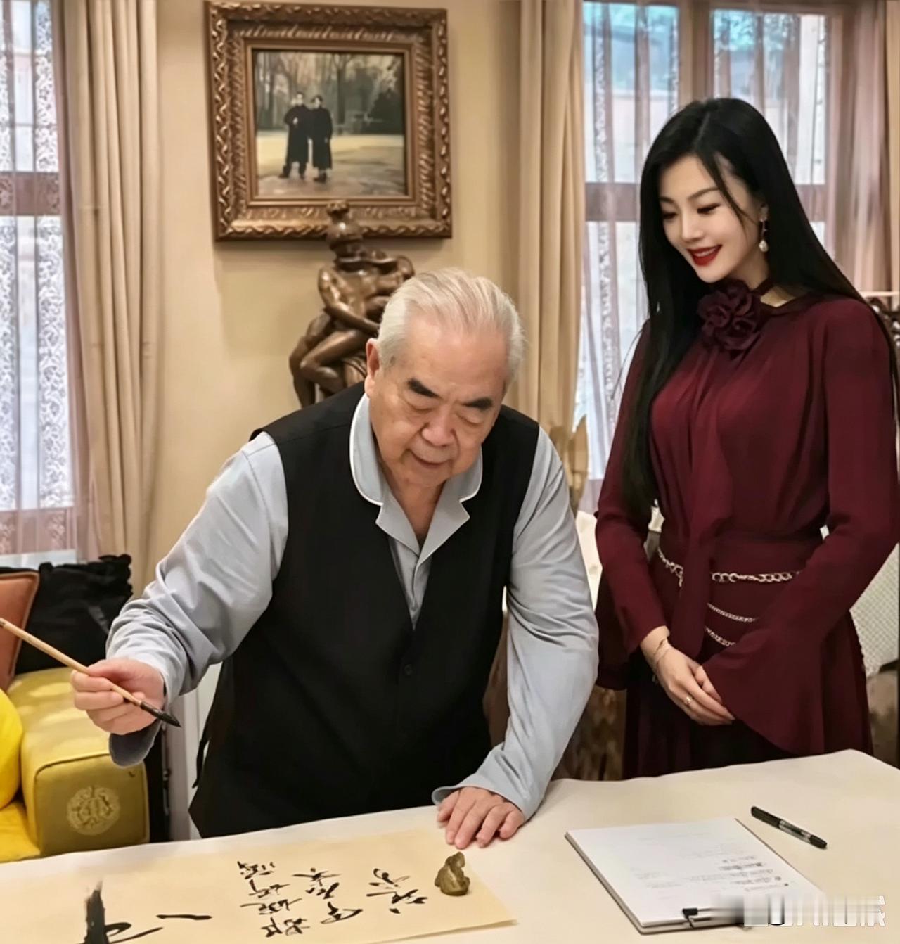 范曾大师正趴书桌上挥毫呢，毛笔甩得飞起，墨点子都快溅到宣纸外头了。旁边站着个美女