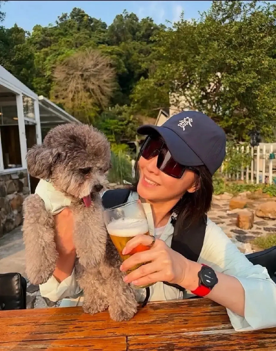 刘嘉玲分享她的灰色泰迪爱犬户外休闲合影，她头戴印有“發”字的深色棒球帽，佩戴大框