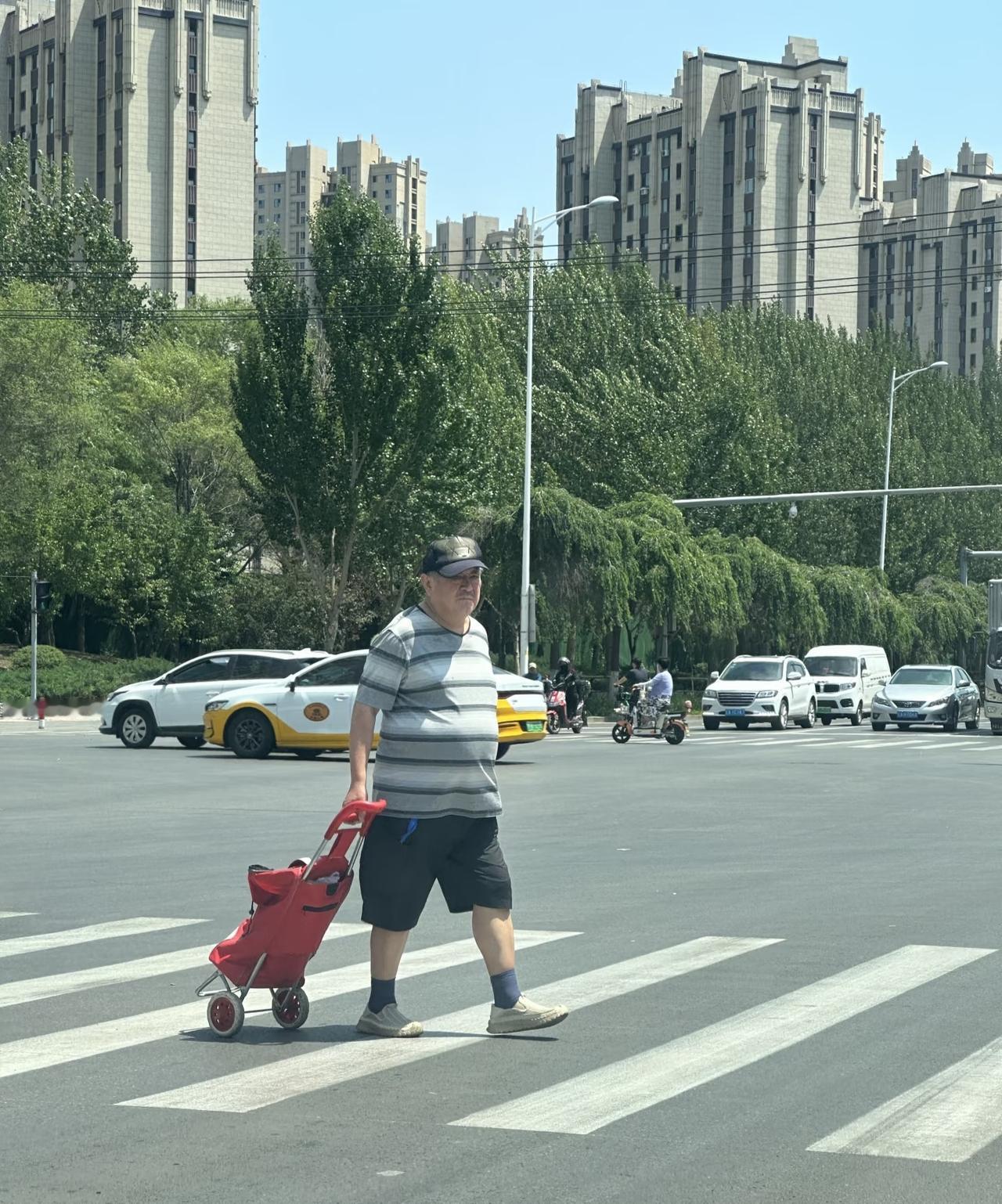 赵本山独自一人过马路，拉着一个红色的折叠购物车，穿着条纹T恤，大短裤，肚