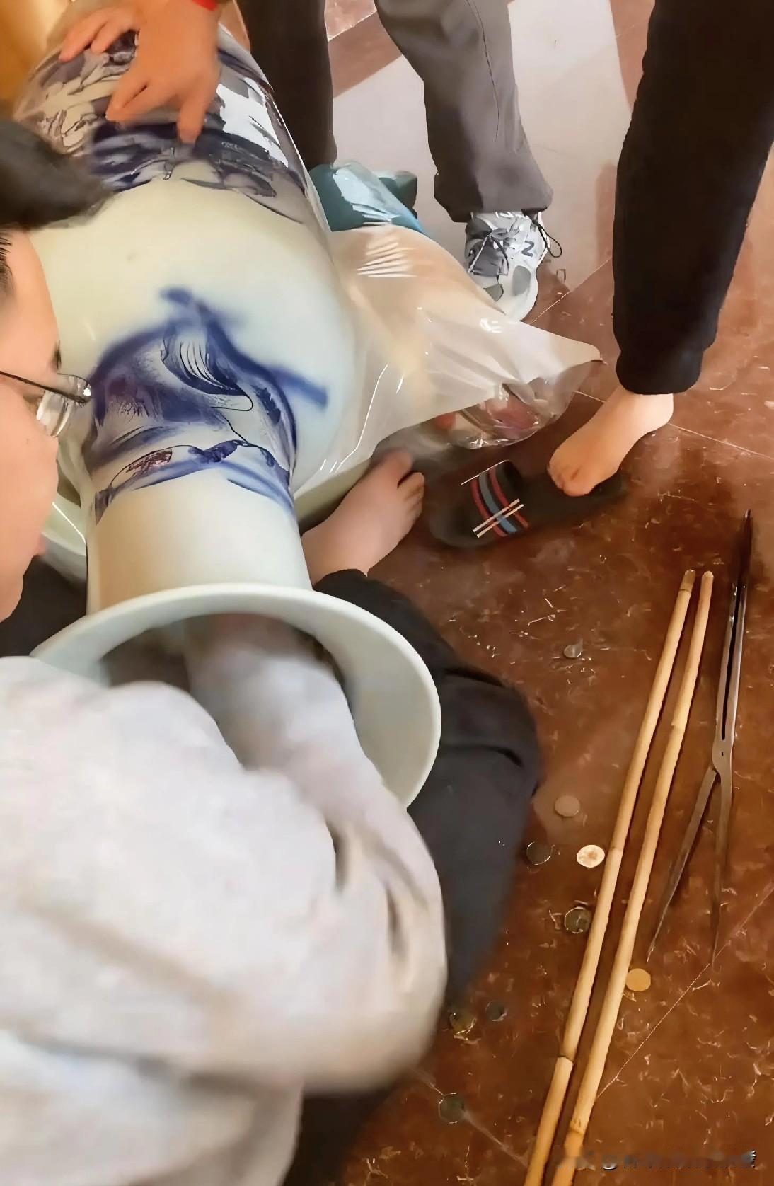 2个人抱起家里装饰用的大花瓶，口朝下，而另外1个人坐在地上，握着花瓶口来回晃动，