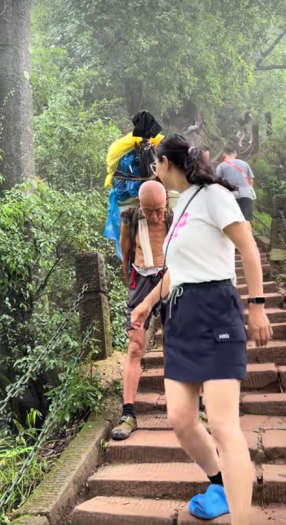 景区的石级山道上，一名游玩的年轻女子，路过一位背山工老人身旁时，回头看了老人一眼