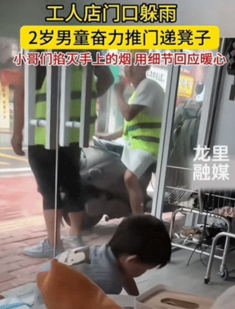 几名工人来到店门口躲雨，店内2岁男童看到后，奋力的推开厚重玻璃门，想把板凳拿给他
