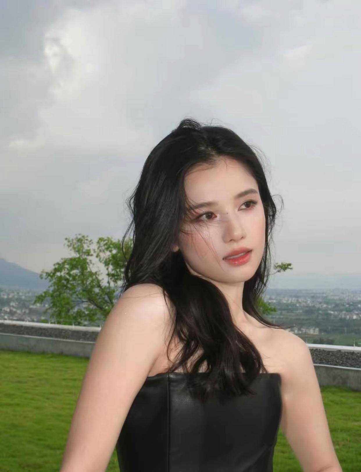 张婧仪身着简约黑色抹胸上衣，乌黑卷发随性散落，眉眼温柔灵动。户外草地与远山作背景，她姿态松弛，时而抬