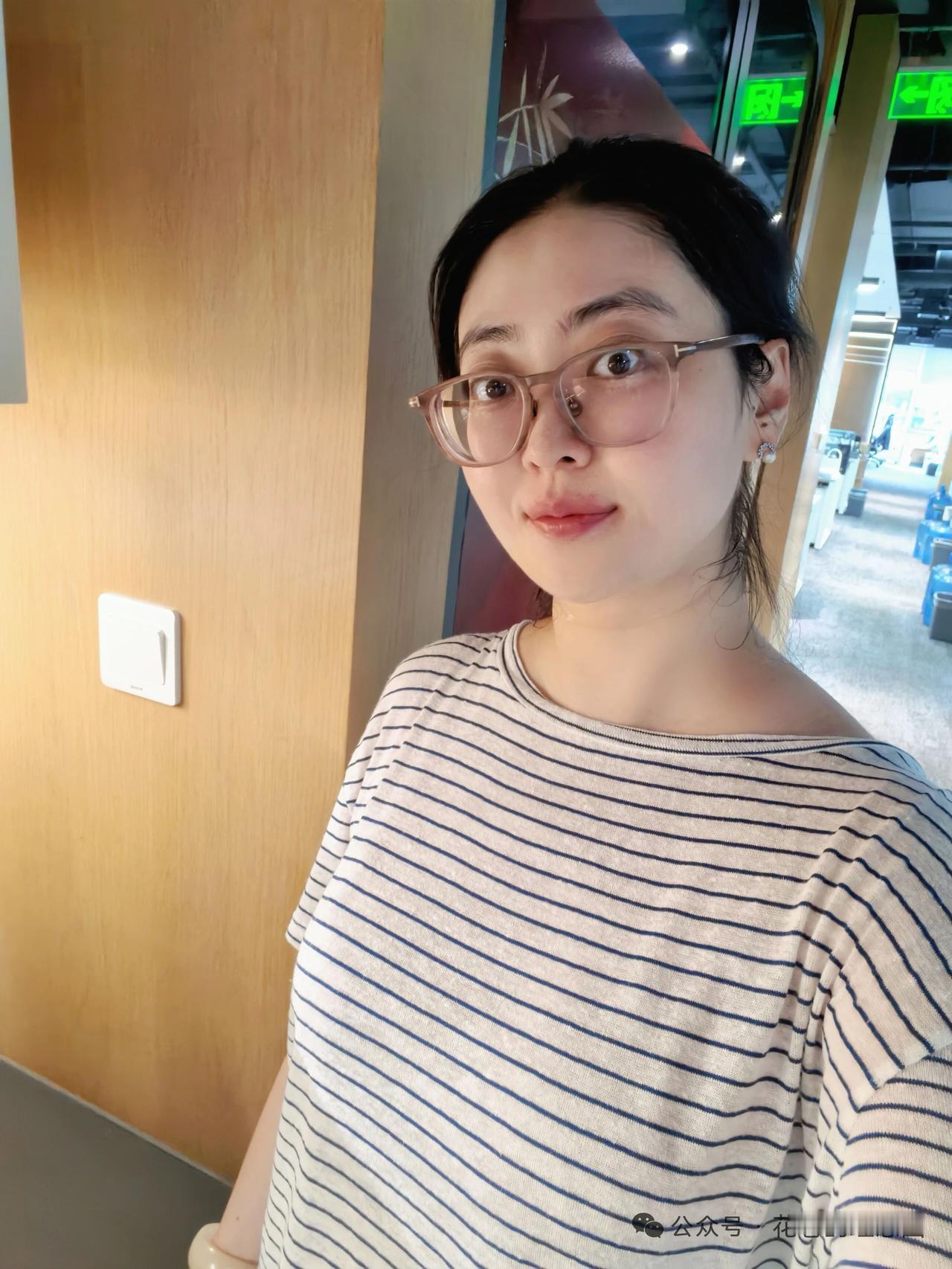 这位姐姐穿着条纹衫，戴眼镜，站在墙边。