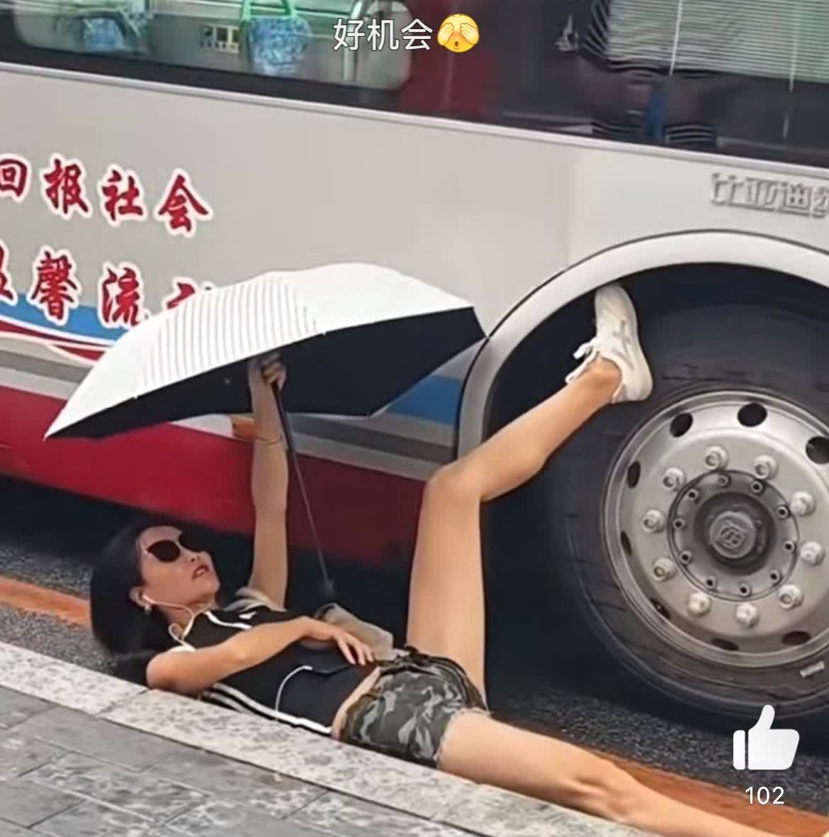 看姿势女子是和公交车杠上了。一只脚搭在公交的轮胎上，人躺在地上，关键是还撑着伞，