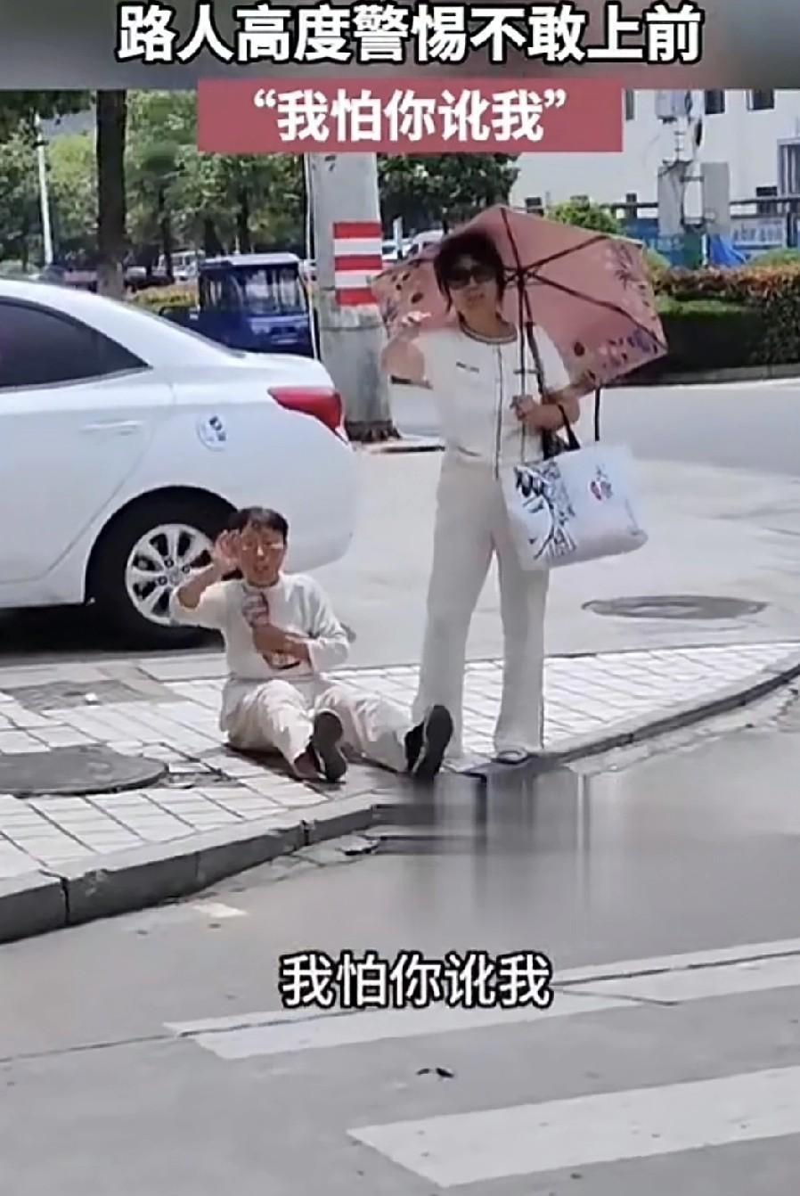 女子的老妈在马路的斑马线前倒地不起，女子打着遮阳伞，手上挎着个袋子，站在一旁。女