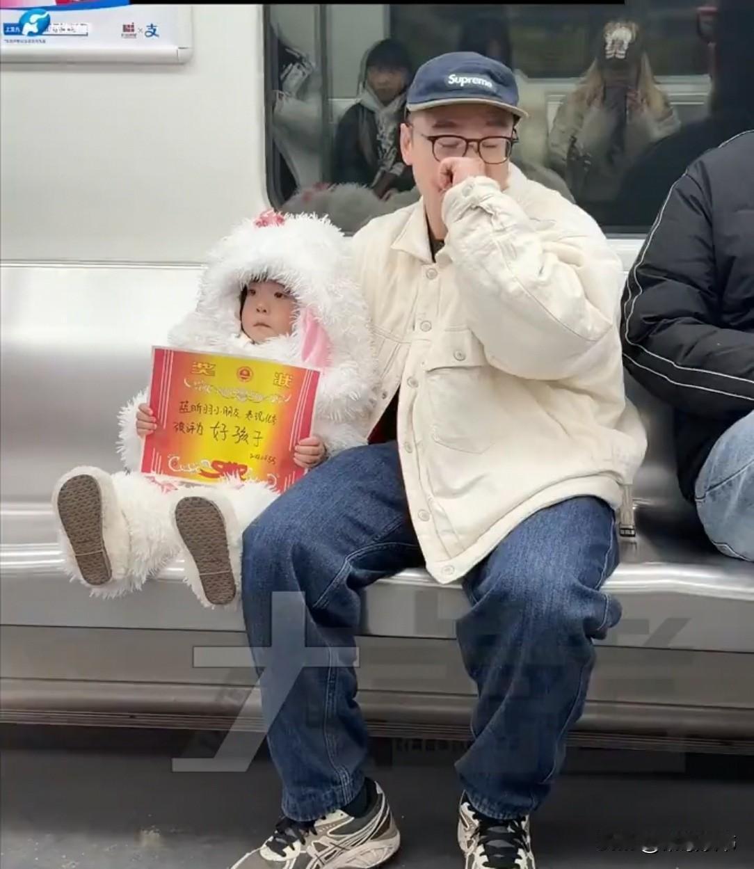 广东一地铁里，男子捂鼻转头，满脸尴尬。身旁娃高举奖状，满脸写着：“快来夸我啊！”