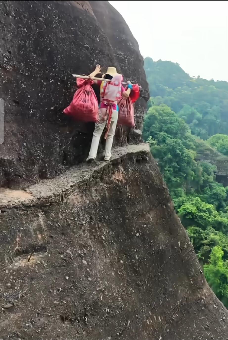 图片中这名妇女肩挑两件东西，后背上还背着一个娃娃，在非常窄的陡峭的大山上行走着！