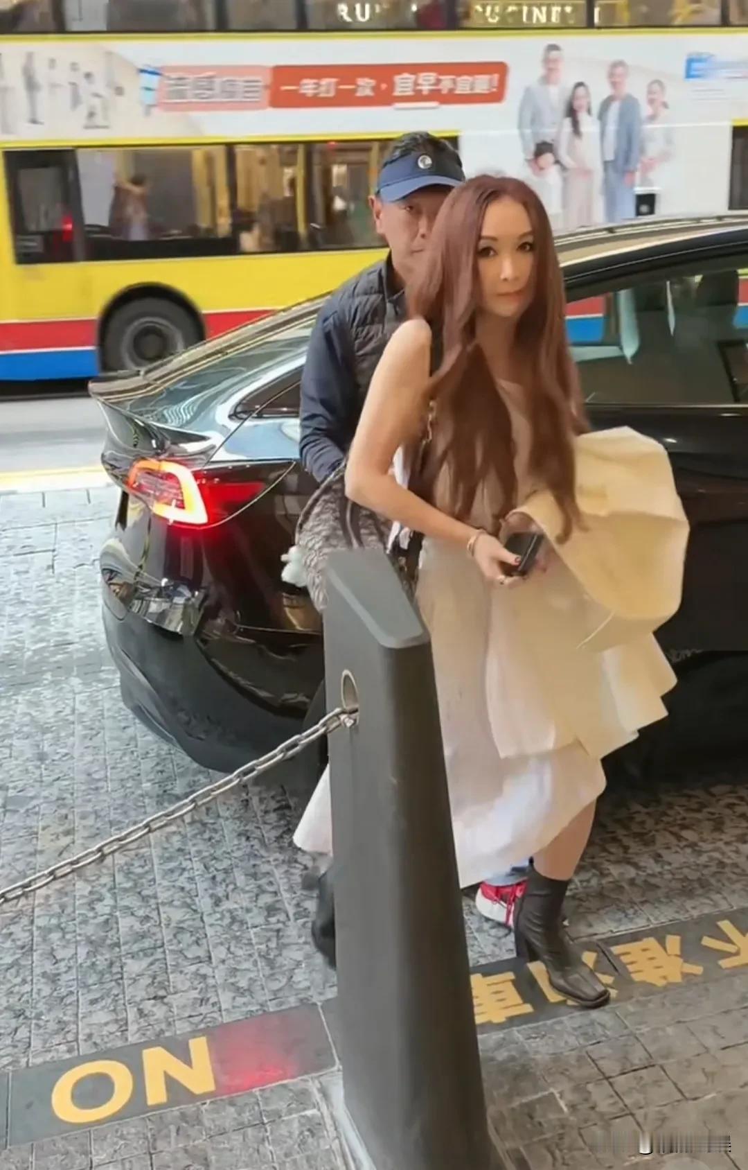 温碧霞和老公在香港街头逛街，一头长发披肩，额头很肿，发际线有点高。她穿着一件露肩