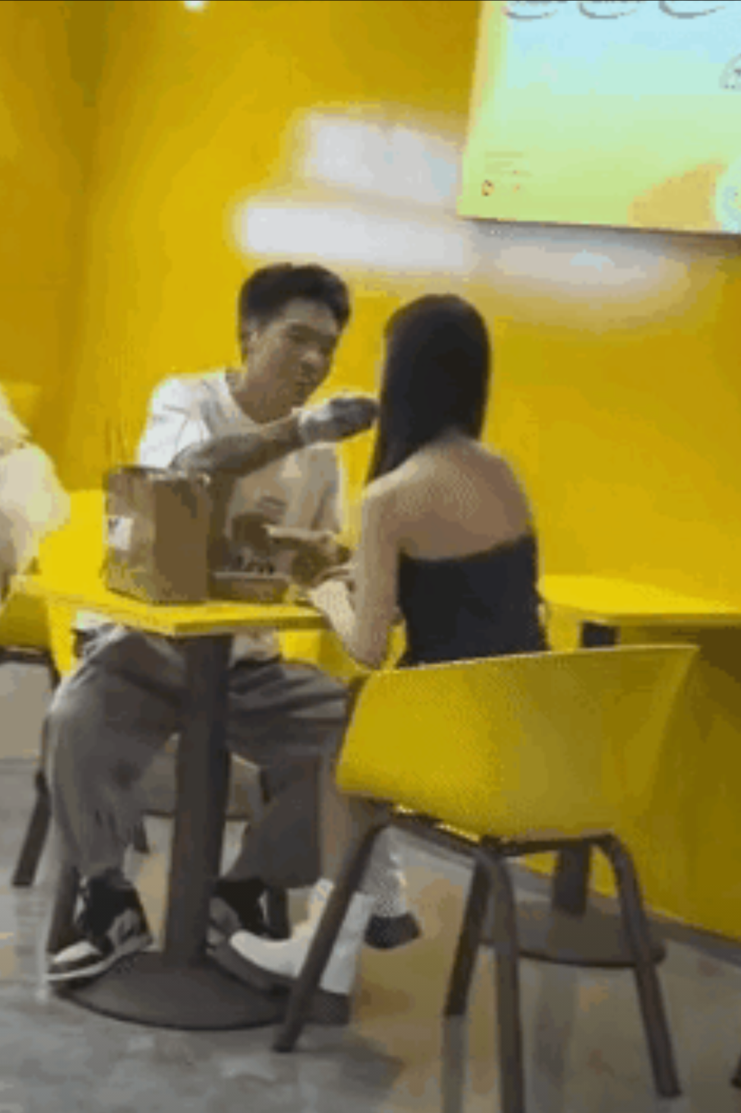 一对情侣去店里吃卤味，男生手上戴上手套，拿着一个鸭翅膀喂给女友吃，脸上洋溢着幸福