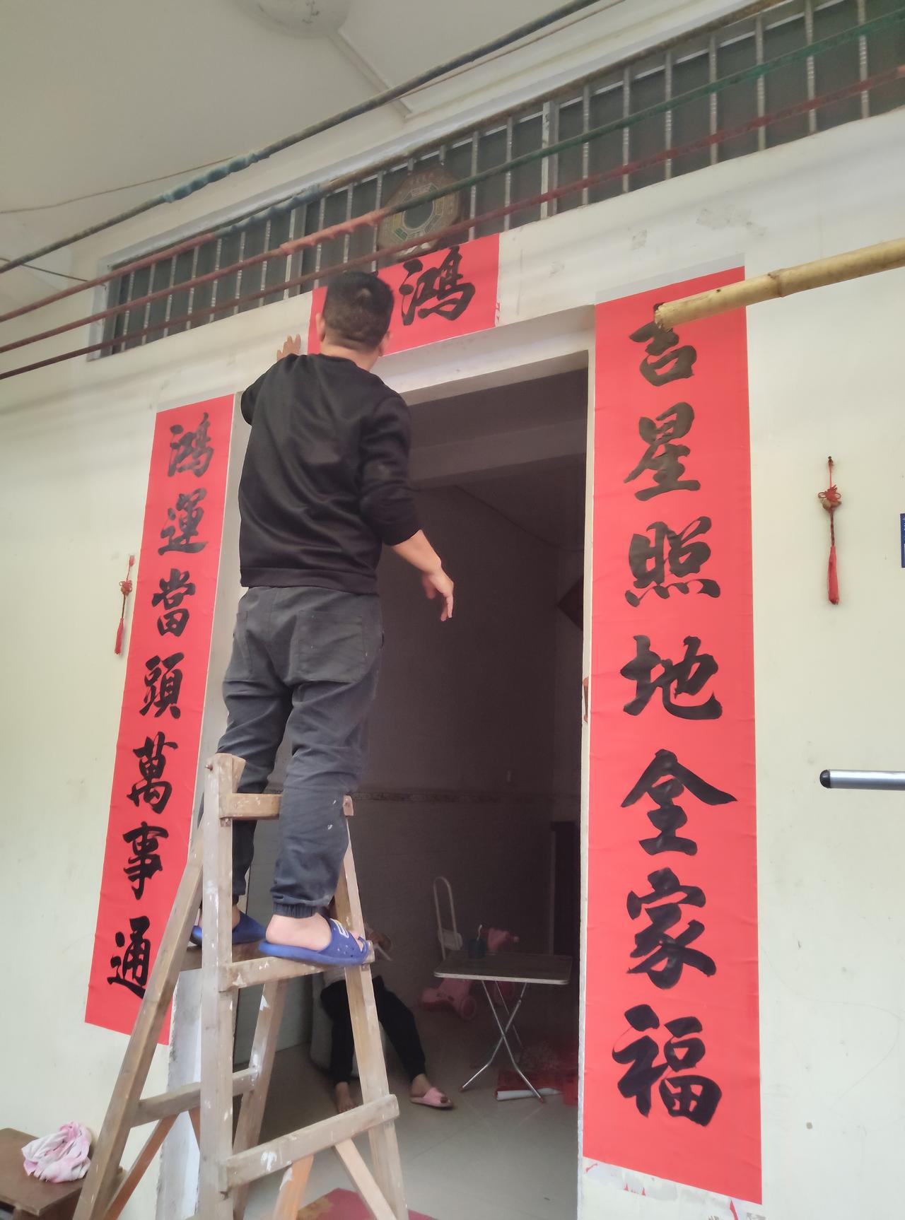 贴春联咯！除夕这天，家里的男主人踩着木梯，赤脚穿着蓝拖鞋就上阵了。左手扶稳门