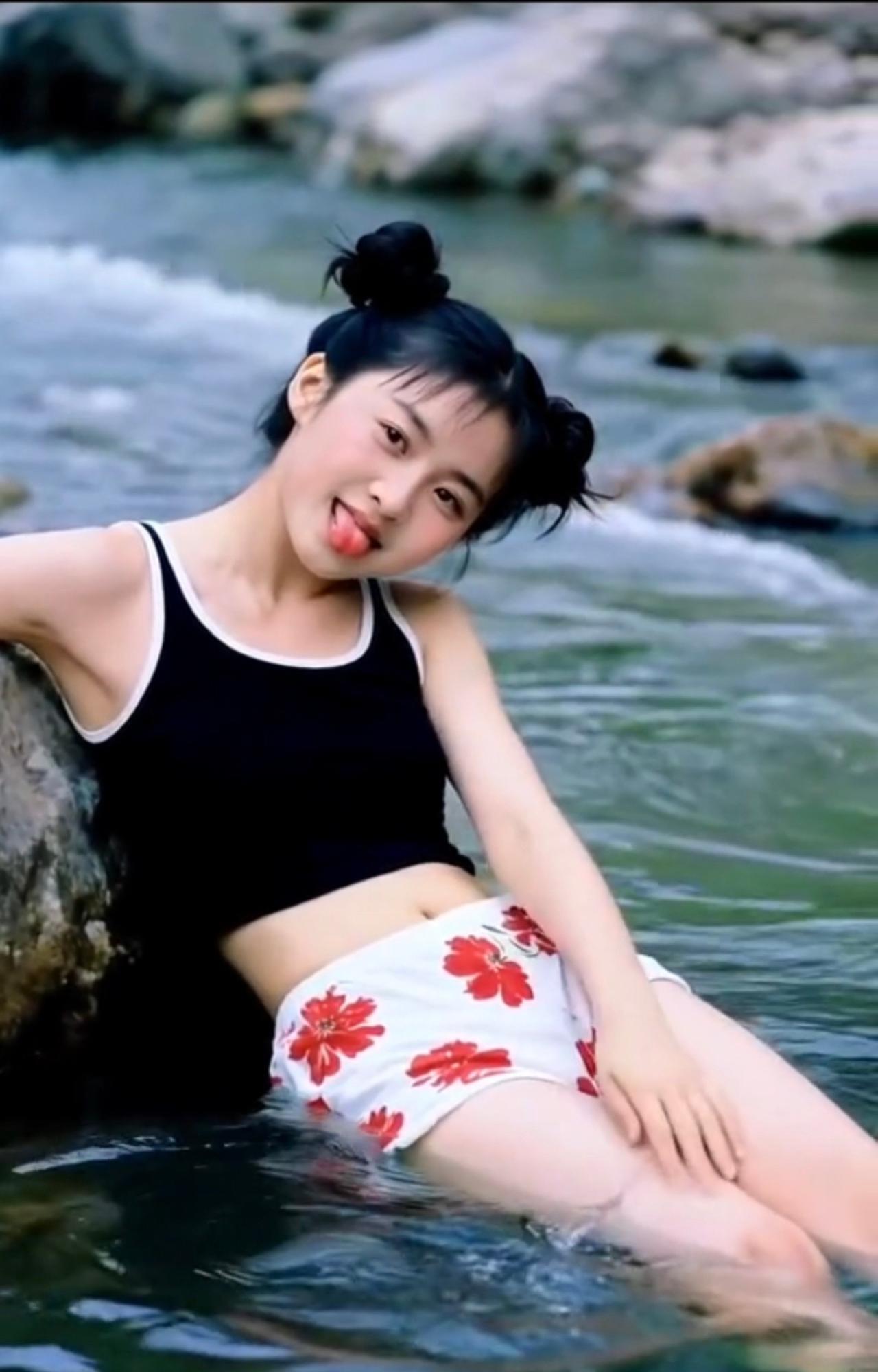 调皮的小妹双丸发髻俏模样，溪边戏水乐无疆。黑衣花裤真灵动，吐舌卖萌趣满塘。