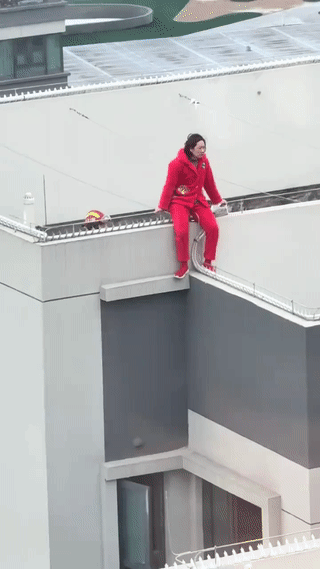 这是一个消防救援现场。一名身穿红色棉服睡衣的女子坐在楼顶上欲跳楼轻生！消防员