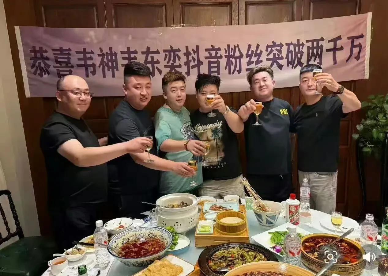 一群人在屋里热热闹闹聚餐。桌上满满当当，全是好吃的菜和酒。背后横幅超醒目，